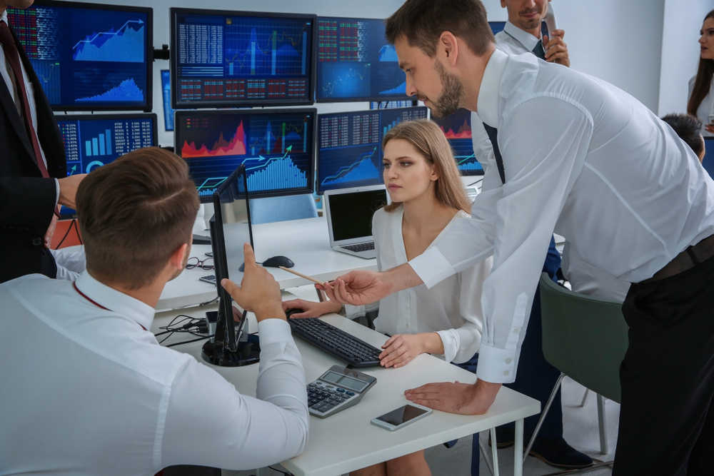 Team of professionals collaboratively reviewing data on multiple monitors displaying charts in a trading room
